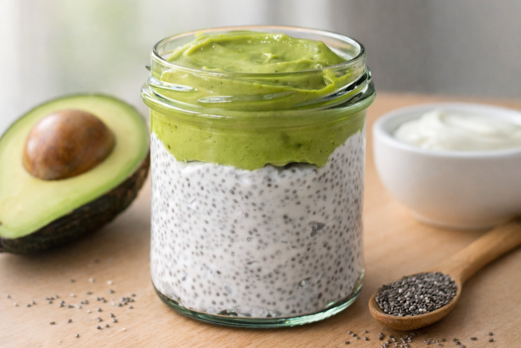 Avokádová pěna na chia pudinku ve skleničce, ozdobená půlkou čerstvého avokáda, miskou jogurtu a lžičkou chia semínek na dřevěném stole.